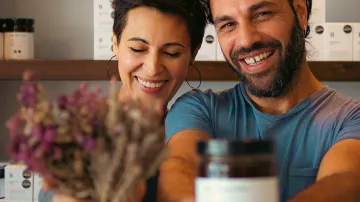 "Chara Mangana and George Athanasiadis, co-founders of Bee Naturalles, smiling and proudly holding a jar of organic honey and a bouquet of dried herbs inside their workshop"